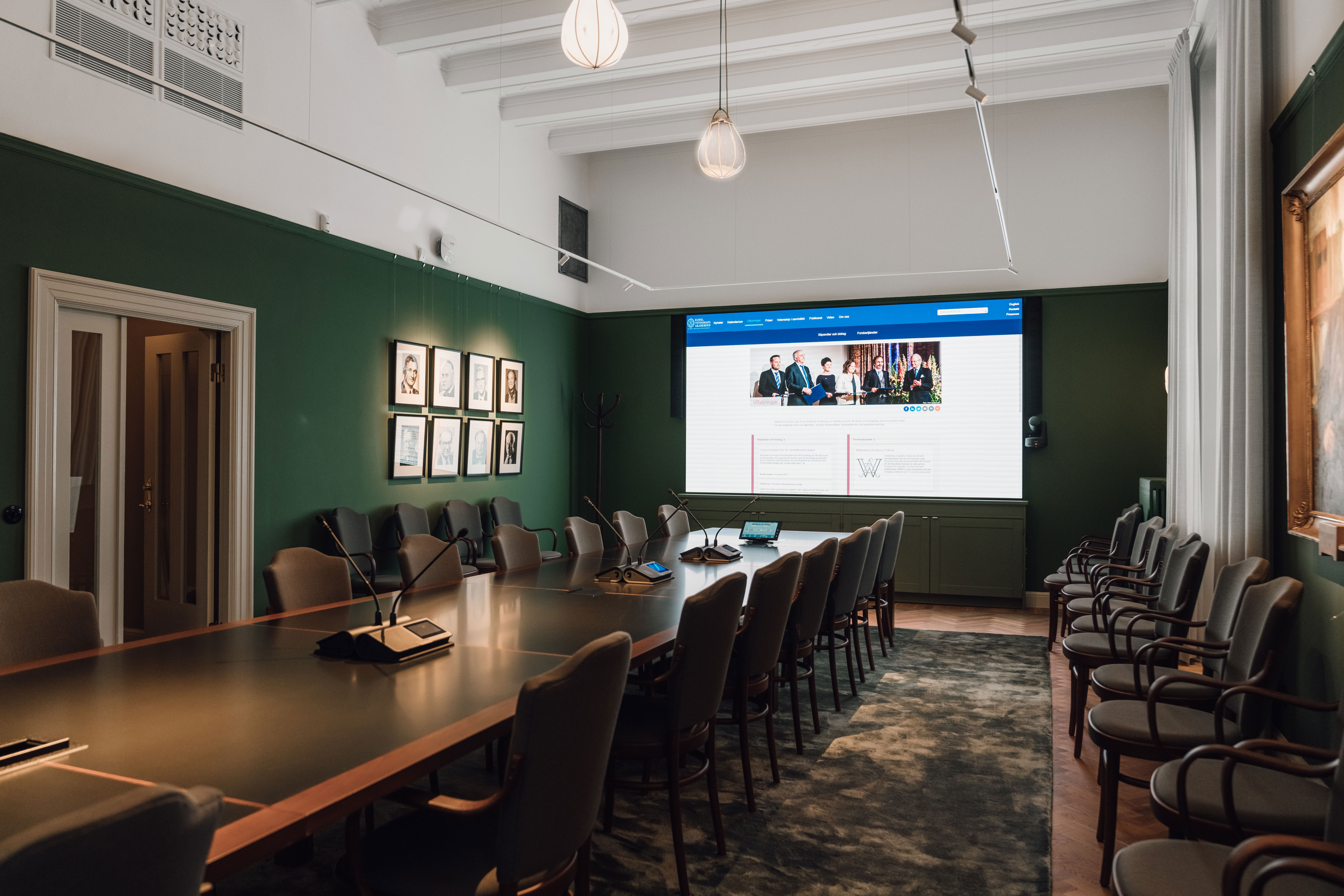 Konferensrum med långbord, mikrofoner och stor skärm som visar mötesteknik, inrett med gröna väggar och porträtt.